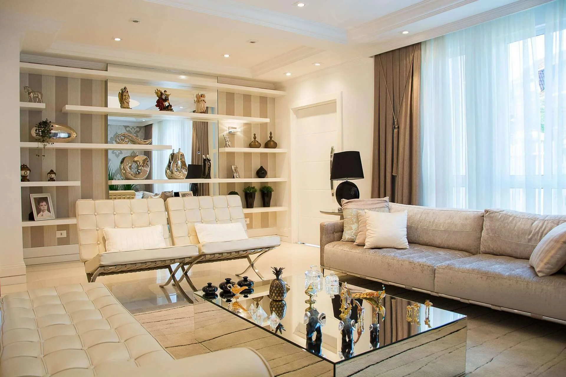 An elegant living room with two white tufted armchairs facing a mirrored coffee table styled with gold and silver decorative objects, a beige sofa along the right wall, and mirrored built-in shelving displaying sculptures and ornaments behind the seating area.