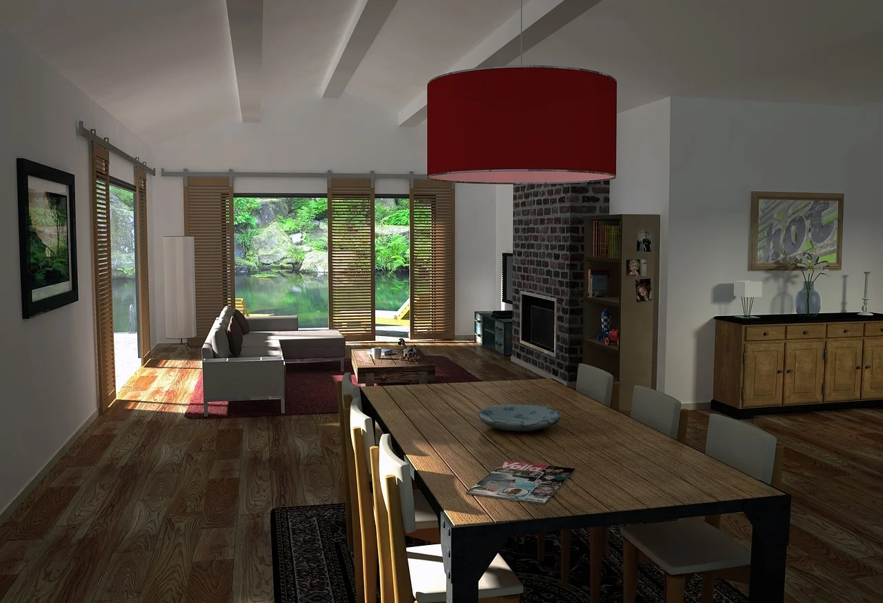 An open-plan living and dining space with a wooden dining table and mixed chairs in the foreground beneath a large red drum pendant light, a white sofa and dark coffee table on a red rug beside a stone fireplace, wooden louvered shutters opening onto a view of a lush green lake, and a wooden sideboard against the white wall on the right.